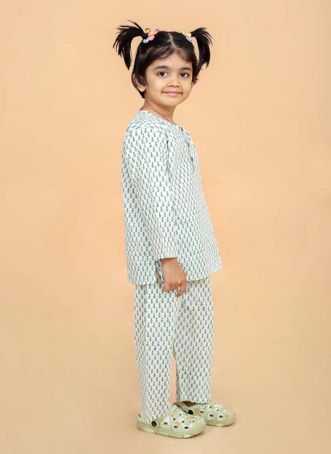 A girl wearing a green booti hand block printed night suit with round neck and half button placket, along with full sleeves and side slits. The set includes matching pajamas with an elasticated waistband.