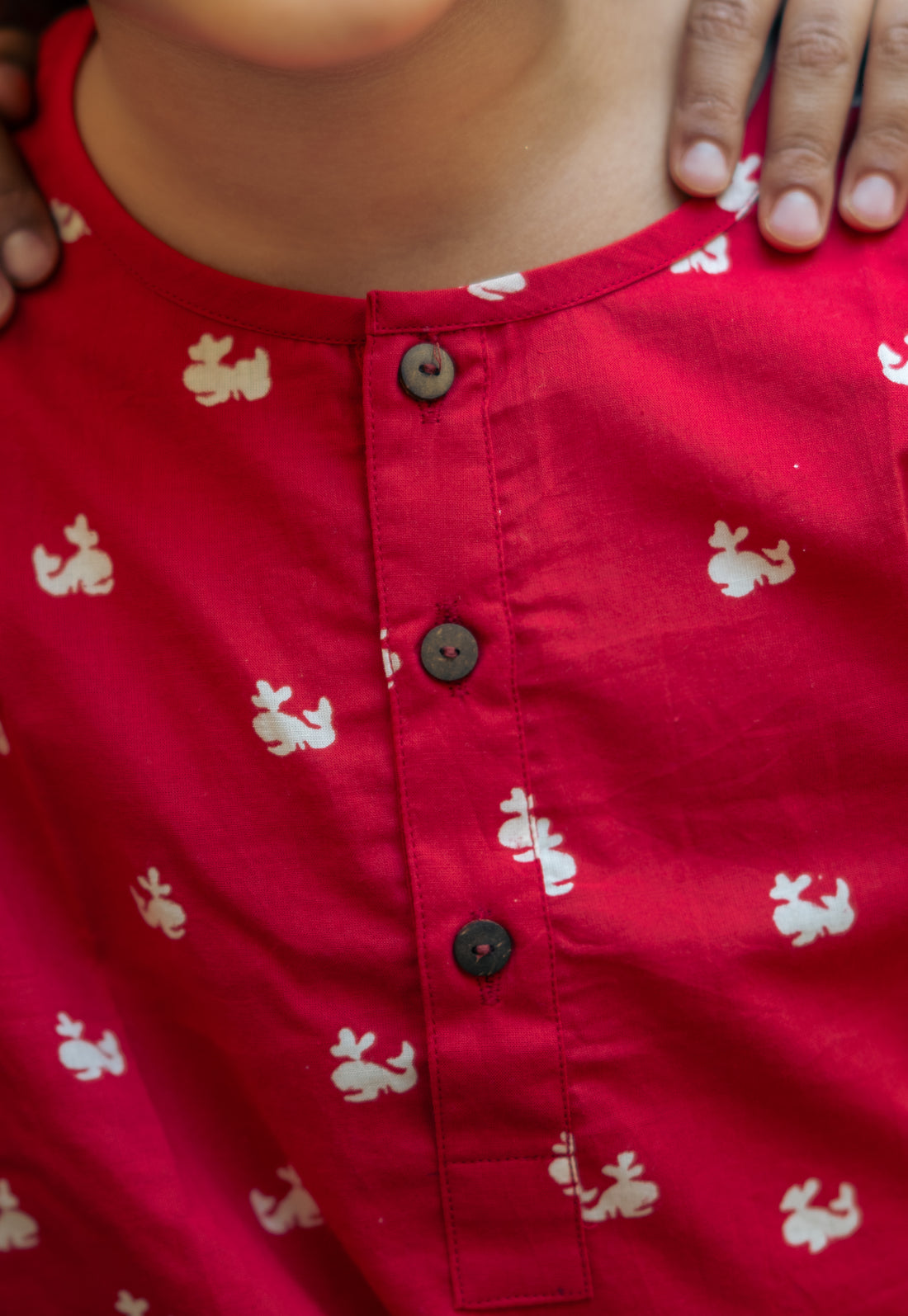 A kid wearing a red night suit with a round neck, half button placket, full sleeves, side slits, and elasticated waistband standing near a potted plant.