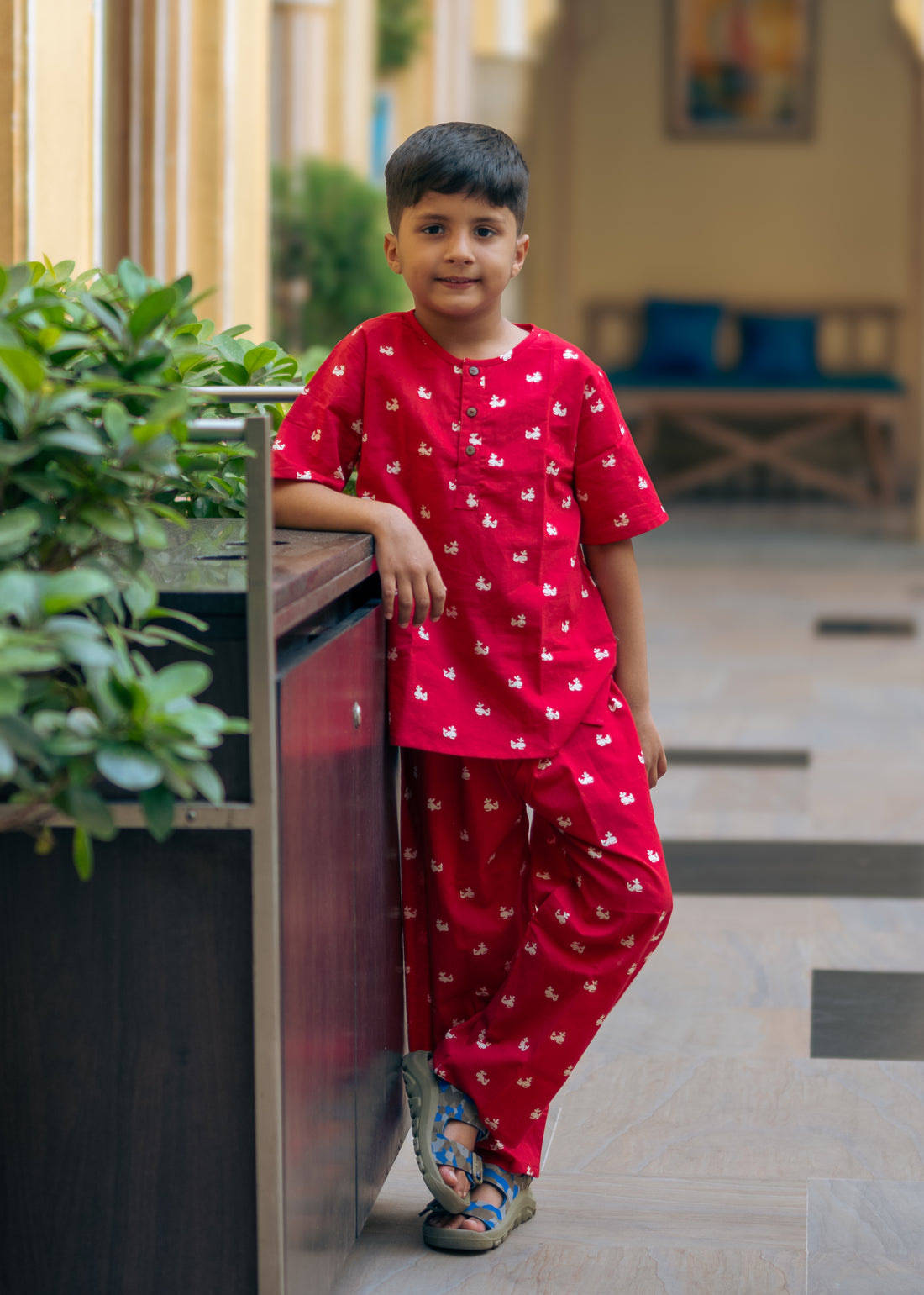A kid wearing a red night suit with a round neck, half button placket, full sleeves, side slits, and elasticated waistband standing near a potted plant.
