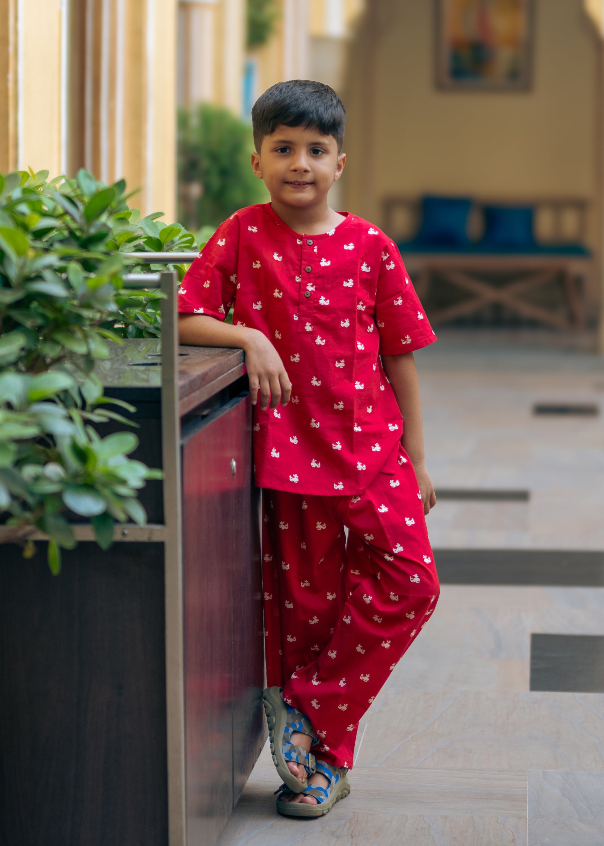 A kid wearing a red night suit with a round neck, half button placket, full sleeves, side slits, and elasticated waistband standing near a potted plant.